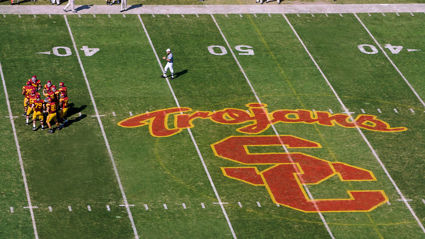 A photo of the USC logo on the football field with the word &quot;Trojans&quot; above it.