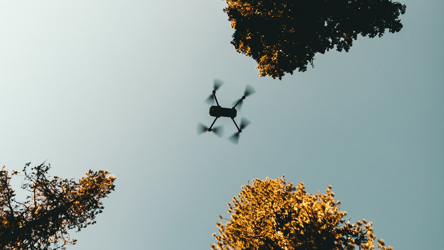 Color photograph looking up at a drone flying in a blue sky.