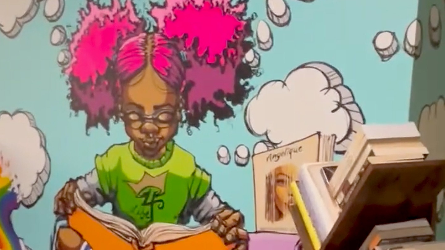 Color photograph of a painted mural featuring a young girl sitting on stacks of books and reading a book.