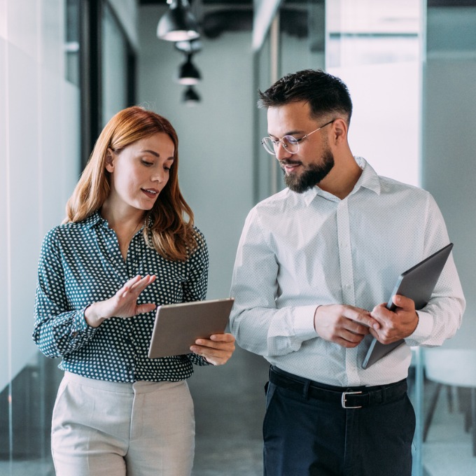 A woman and a man walking in an office looking at an ipad