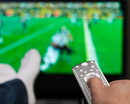 Color photograph of a person watching football on TV with remote control in hand