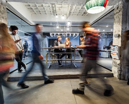 A man and a woman sit and talk at a table while people walk by them.