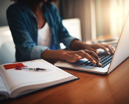 a woman's hands typing on a computer next to an open notebook.