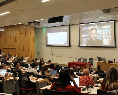 Conference attendees sit in a lecture hall and listen to a virtual presentation from Sal Khan.