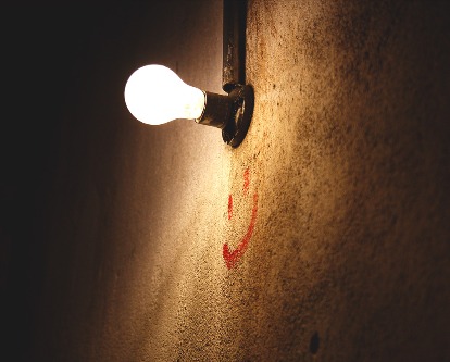Color photograph of a lit lightbulb above a smiley face painted on a wall.