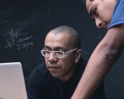 Color photograph of two people in a classroom looking at a laptop.