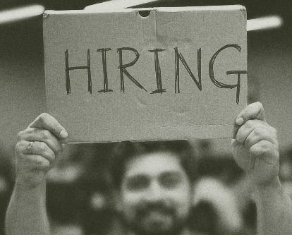 Olive duotone image of a male-presenting person holding up a handwritten sign reading &quot;Hiring&quot;.
